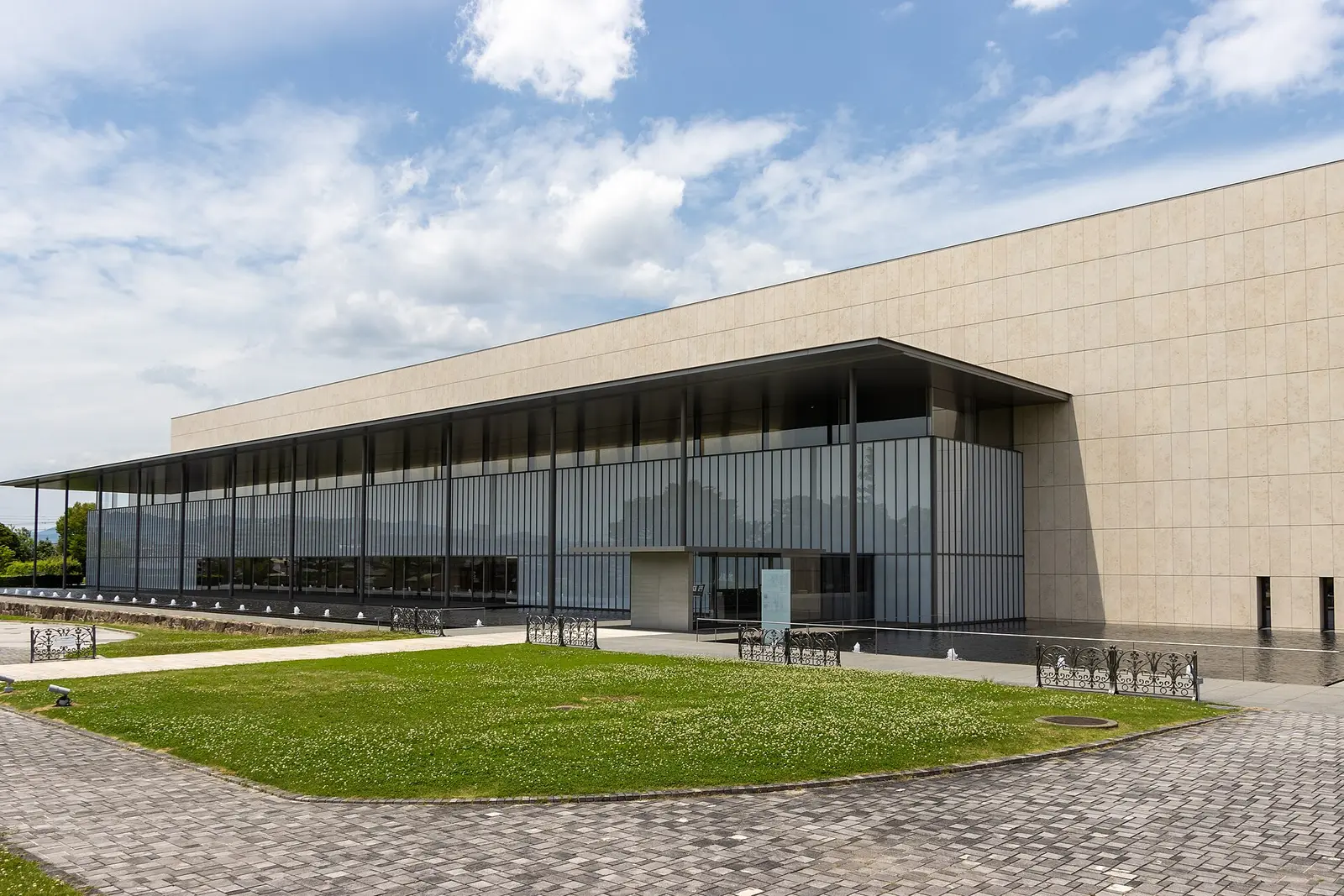 Glass fronted Heisei Chishinkan Wing of the Kyoto National Museum beneath a stone clad block, seen across a lawn and paved forecourt
