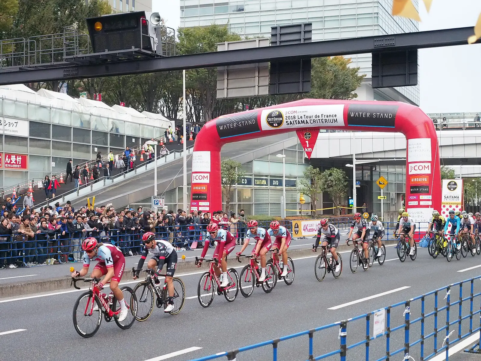 Peloton riding past an inflatable arch and spectators behind barriers at the 2018 Tour de France Saitama Criterium near Saitama Shintoshin Station